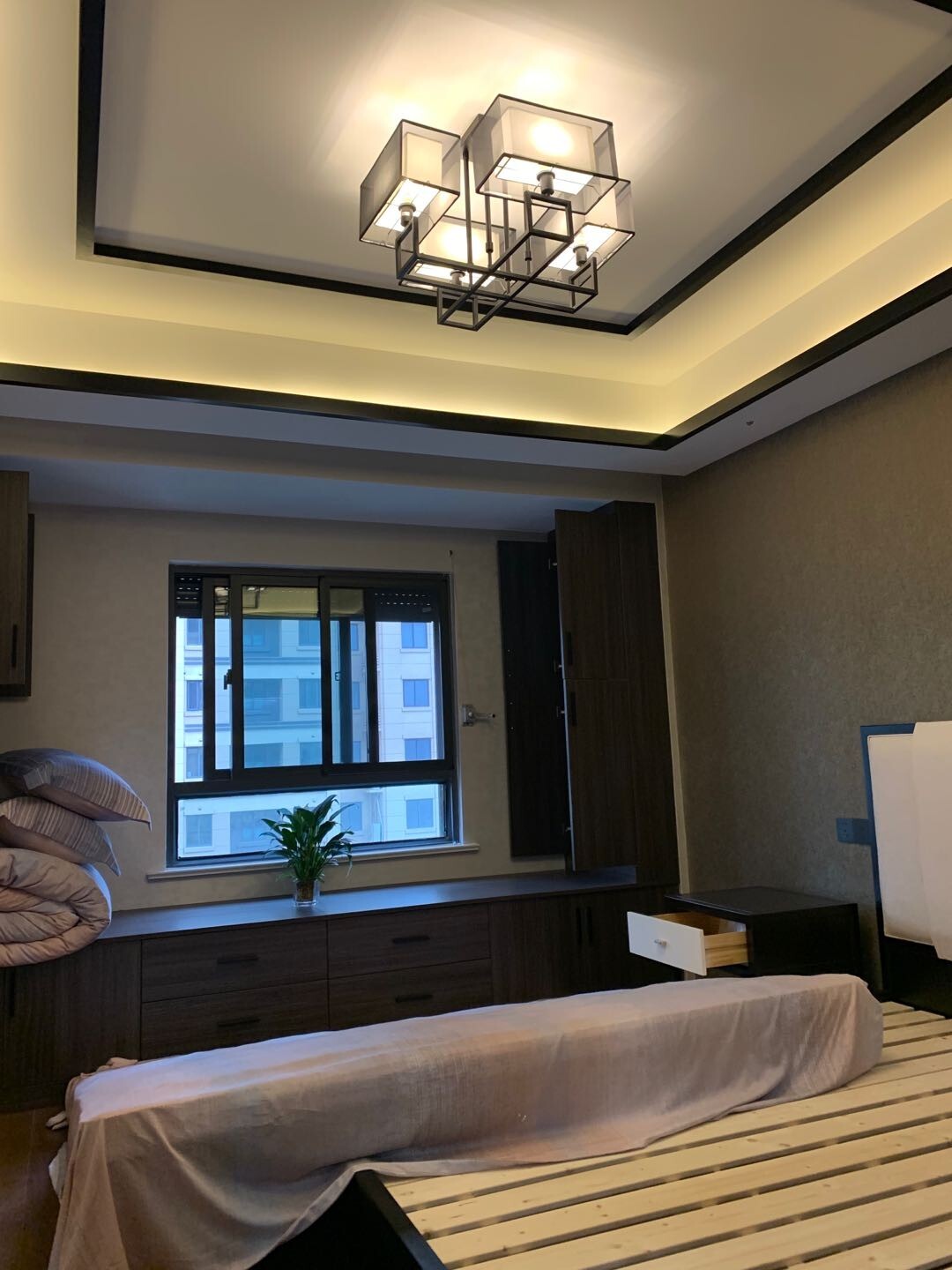 新中式两房卧室,深木衣柜、窗下抽屉柜、米灰墙面、深色顶面线条与几何灯具组成休息区